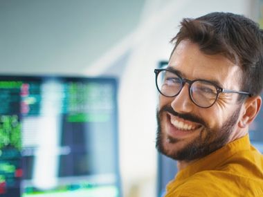 Stellenagebot ansehen: IT-Mitarbeiter:in (m/w/d) bei PSW GROUP Lächelnder Mann mit Brille an einem Arbeitsplatz mit mehreren Monitoren.